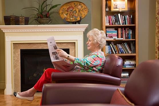 An elderly woman reads a newspaper in a cozy living room setting.