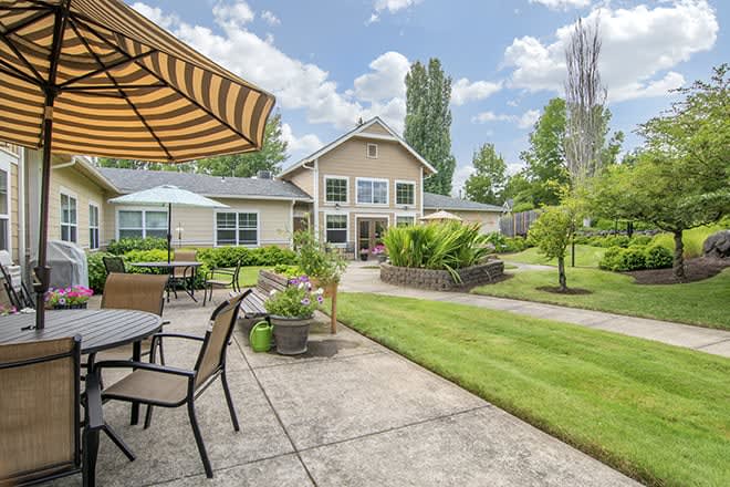 A beautiful backyard with a well-maintained lawn, patio furniture under a striped umbrella, and lush greenery surrounding a modern house.