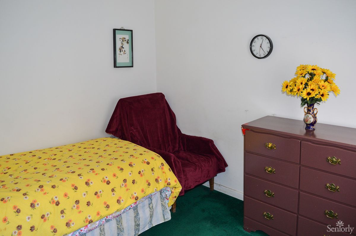 A simple bedroom with a single bed, a small dresser, and a chair, featuring minimal decor including a wall clock.