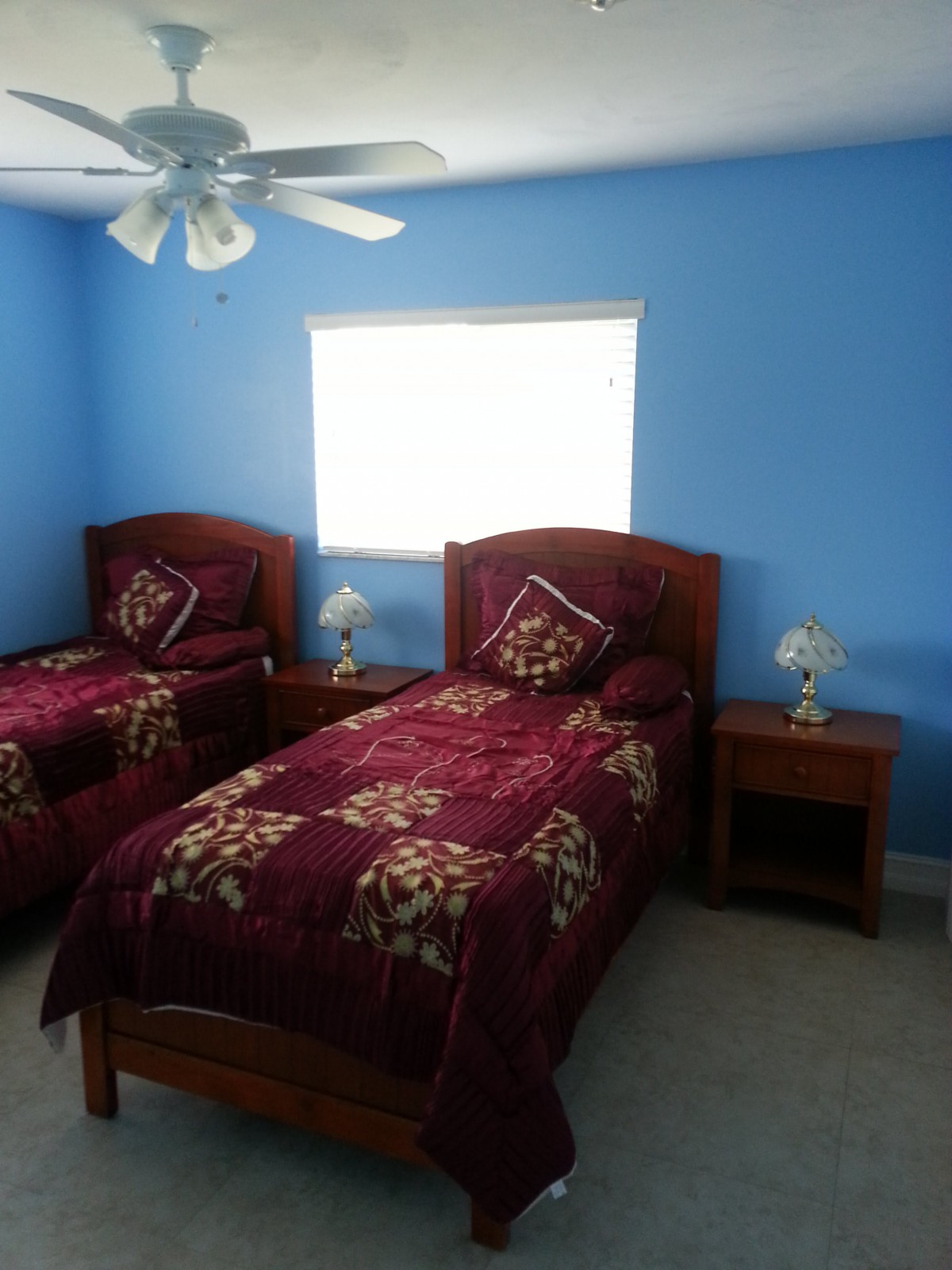 A cozy bedroom with two twin beds and blue walls, featuring nightstands and table lamps under a ceiling fan.