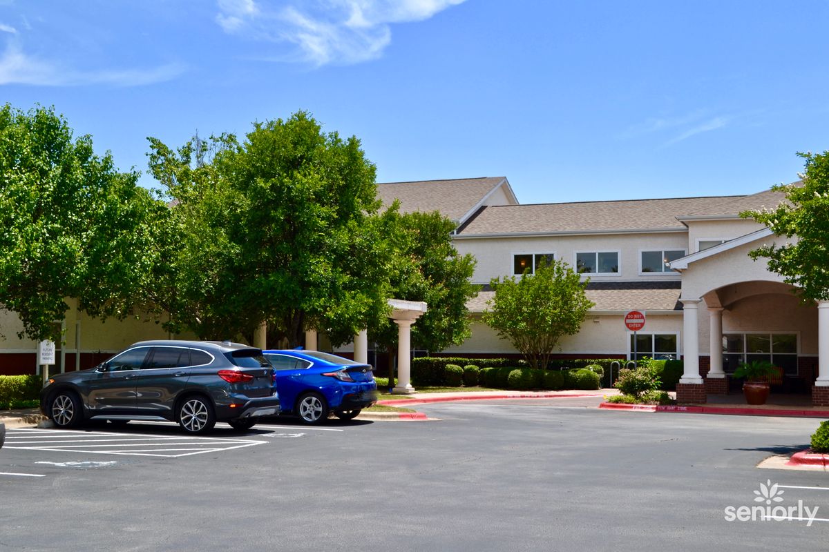 A sunny day view of a parking lot with several cars in front of a building surrounded by trees.