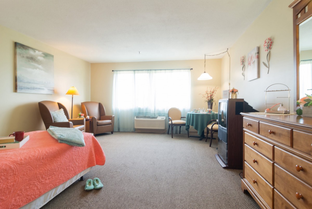 A cozy bedroom with warm lighting, featuring a bed, armchair, and a small dining area.