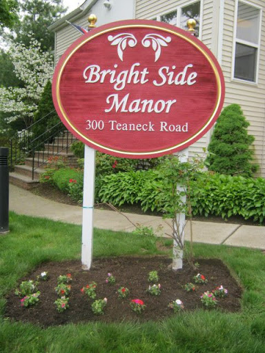The sign for Bright Side Manor, located at 300 Teaneck Road, surrounded by a well-maintained garden.