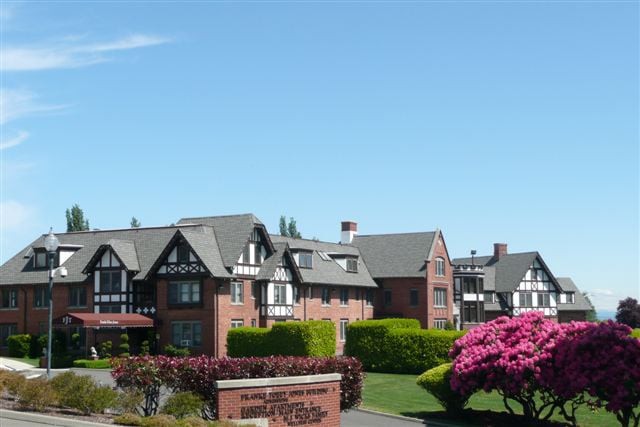 A scenic view of a residential brick building with Tudor-style architecture, surrounded by lush greenery and flowering bushes under a clear blue sky.