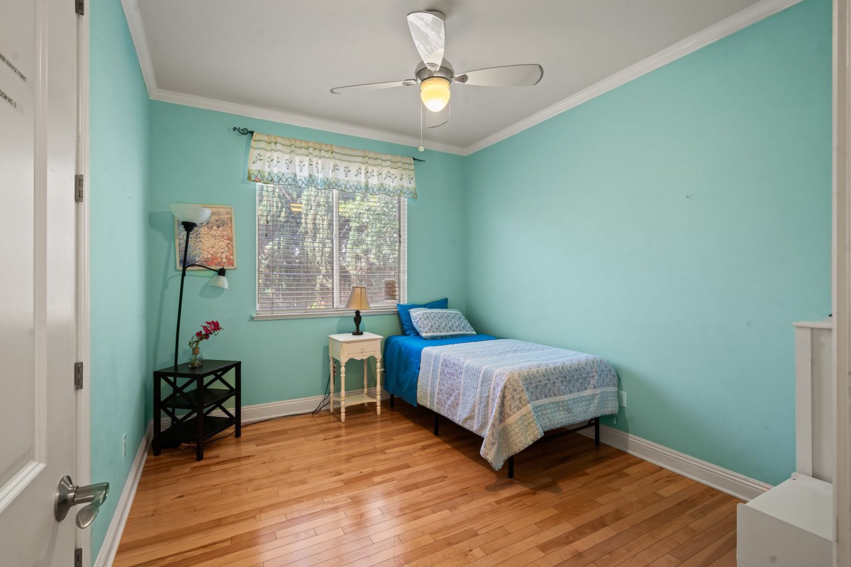 A simple bedroom with a single bed and wooden flooring, highlighted by natural light from the window and adorned with a ceiling fan.