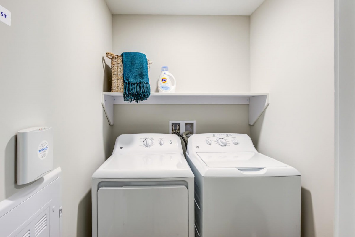 A small laundry room with a washing machine, dryer, and a shelf containing towels and detergent.