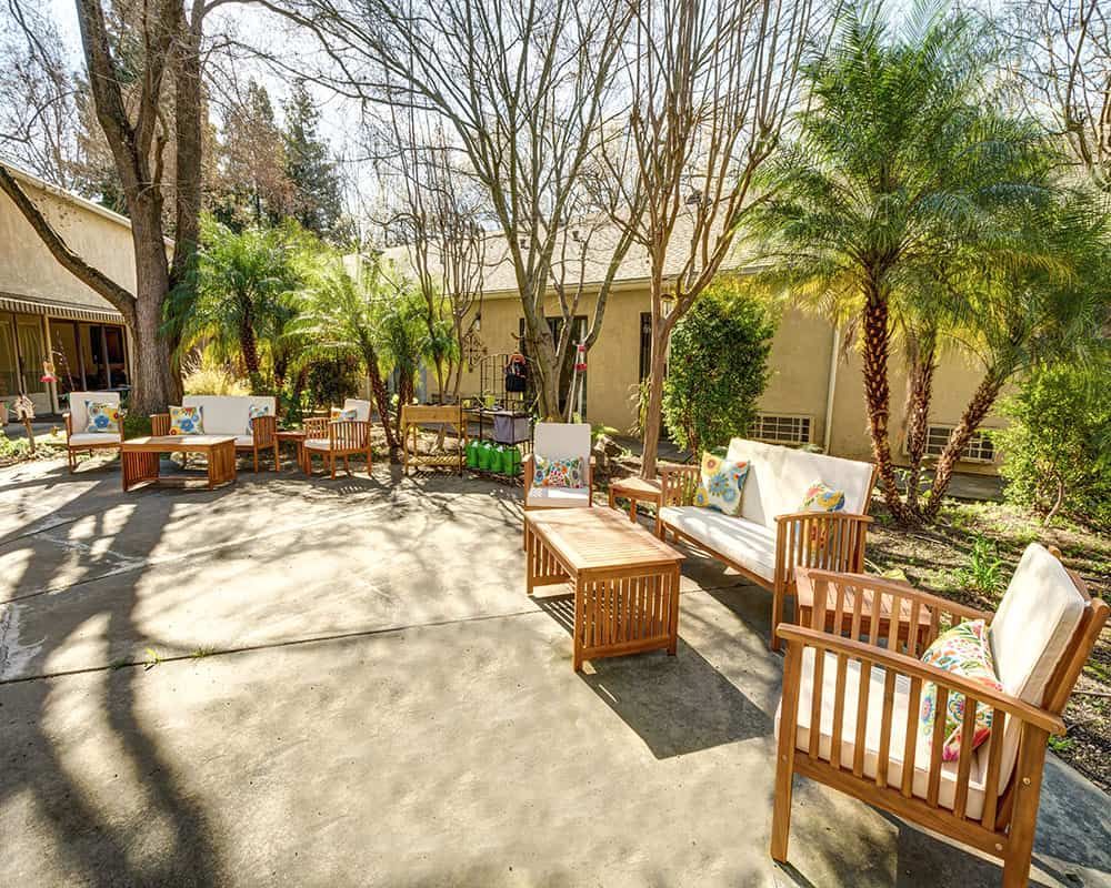 A serene outdoor patio with wooden furniture surrounded by lush greenery and sunlight.