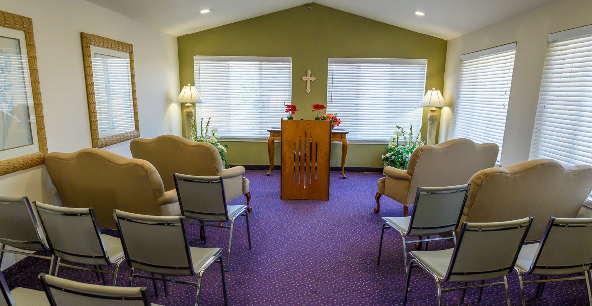 A small chapel interior with seating, plants, and an altar under a cross.