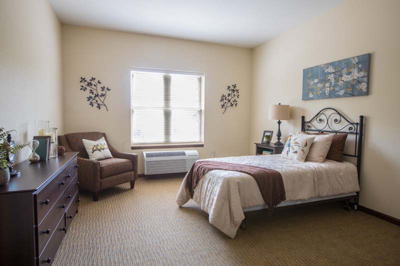 A cozy bedroom with a single bed, armchair, and decorative elements surrounding a bright window.