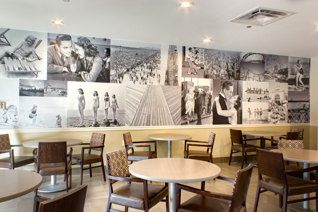 A cozy cafe interior featuring black and white photographs on the wall, with empty tables and chairs.