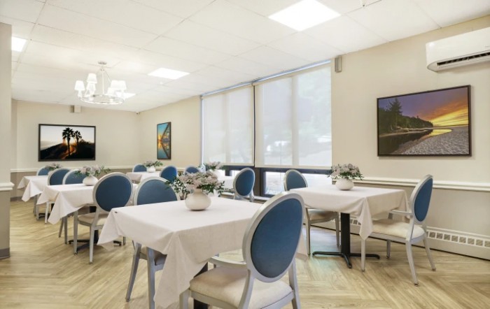 A neatly arranged dining area with tables and blue-cushioned chairs, decorated with potted plants and artwork on the walls.