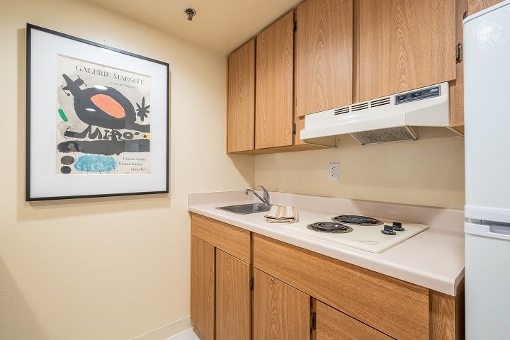 A compact kitchenette with wooden cabinets, a small sink, and a stovetop, accented by a framed poster on the wall.