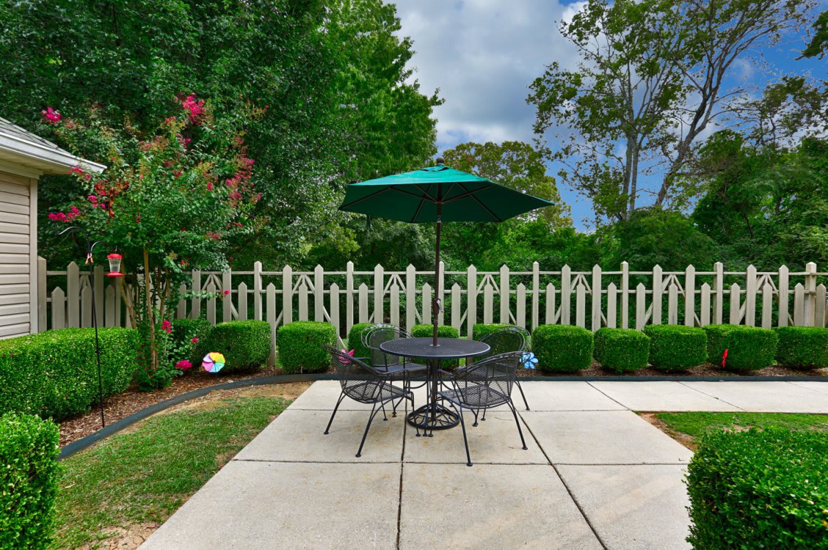 A peaceful patio area with a table and chairs under a green umbrella, surrounded by a garden and a picket fence.