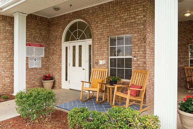 A cozy porch with wooden chairs and potted plants at the entrance of a brick house.