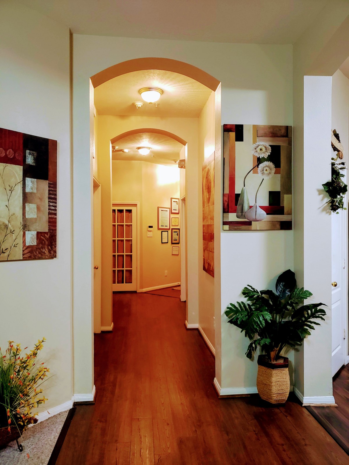 A warmly lit hallway adorned with paintings and plants, featuring a wooden floor leading to a door.