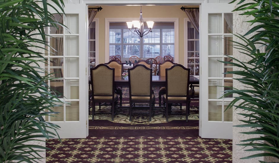 A dining room with a set of chairs around a long table, illuminated by a chandelier, viewed through open French doors.