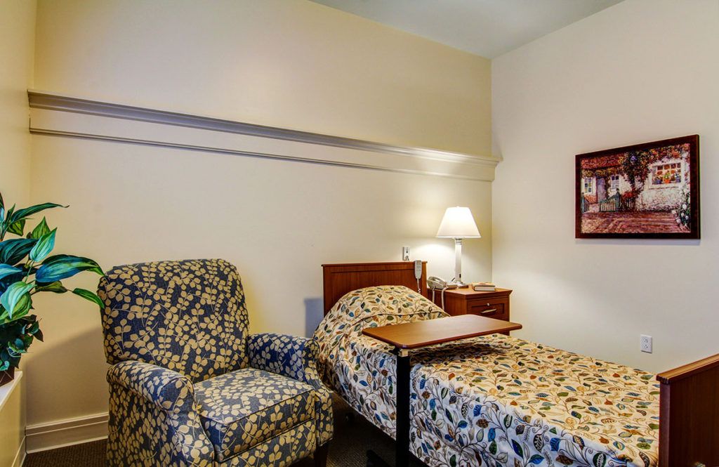 A cozy bedroom with a hospital-style bed and an armchair, softly lit by a table lamp.