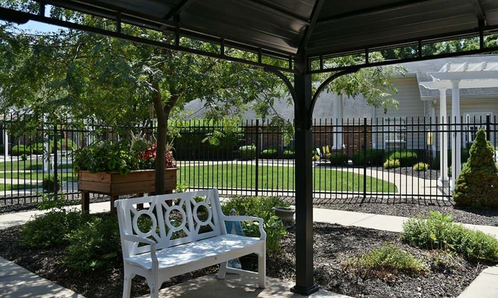 A peaceful garden scene with a bench under a gazebo, surrounded by greenery and enclosed by a fence.