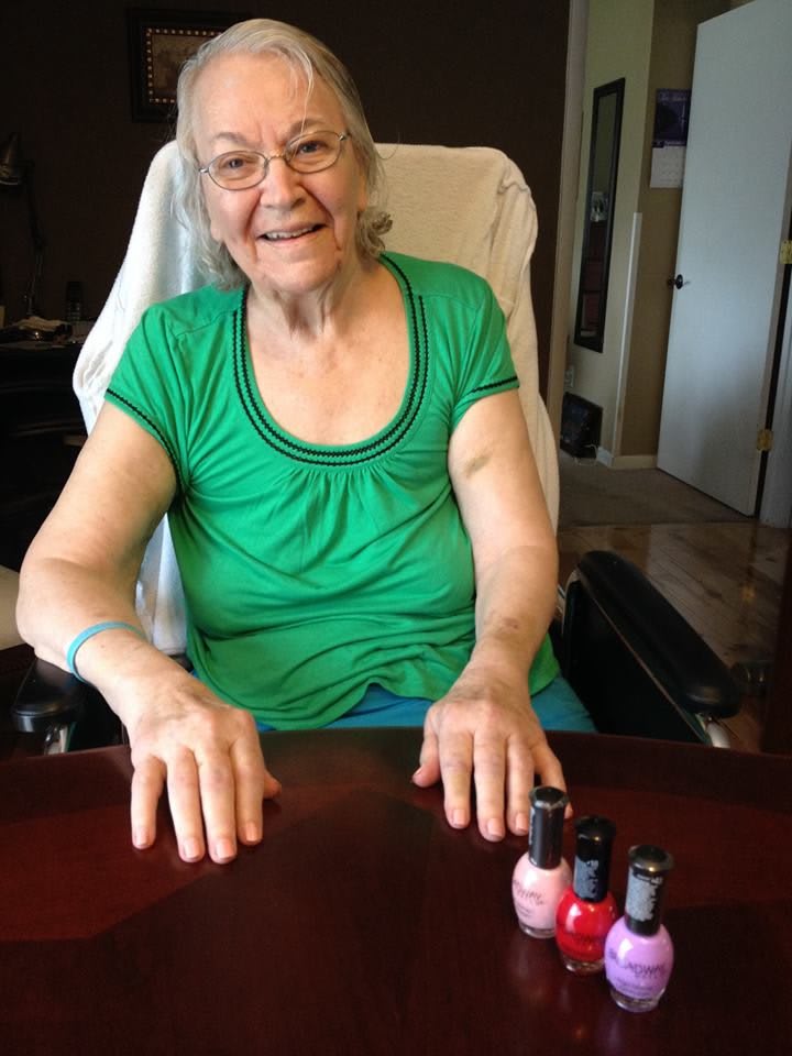 Elderly woman smiling while displaying nail polish on a table.