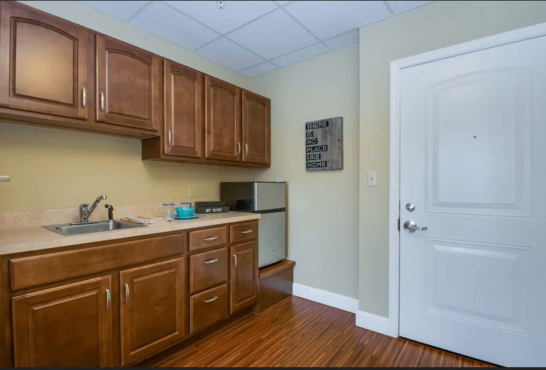 A small kitchen area with wooden cabinets, a sink, compact fridge, and hardwood flooring.