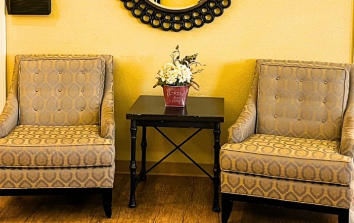 A cozy seating area with two upholstered chairs and a small table with a flower arrangement, set against a yellow wall featuring a decorative mirror.