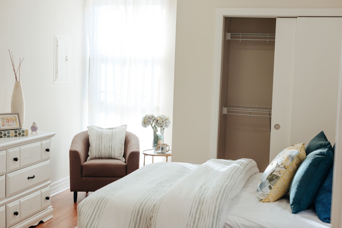 A minimalist bedroom with a bed, an armchair, and a vase on a side table near a window.