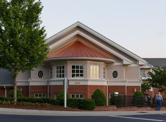A suburban building with a mix of brick and siding, featuring large windows and surrounded by greenery.
