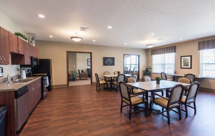 A spacious dining area with wooden flooring, tables, and chairs, well-lit by ceiling lights and natural light from large windows.