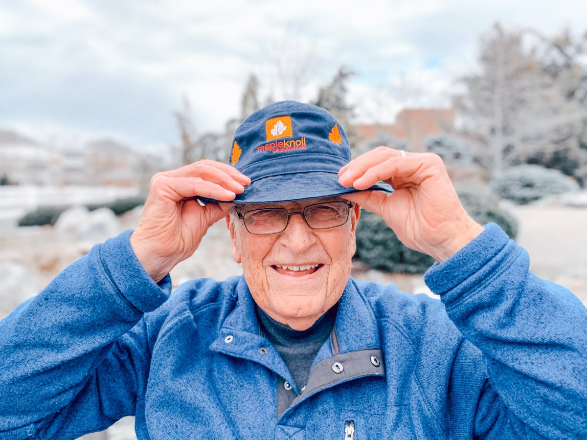 An elderly man smiling and adjusting his cap while standing outdoors.