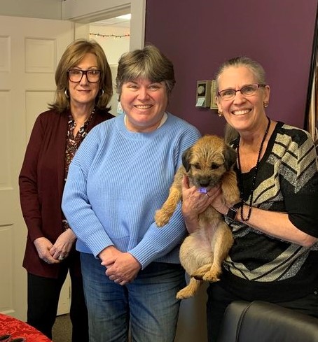 Three women pose together with a small dog in a cozy indoor setting.