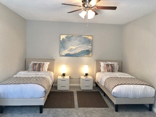 A modern bedroom featuring twin beds with neutral-toned bedding, complemented by a ceiling fan and abstract artwork on the wall.