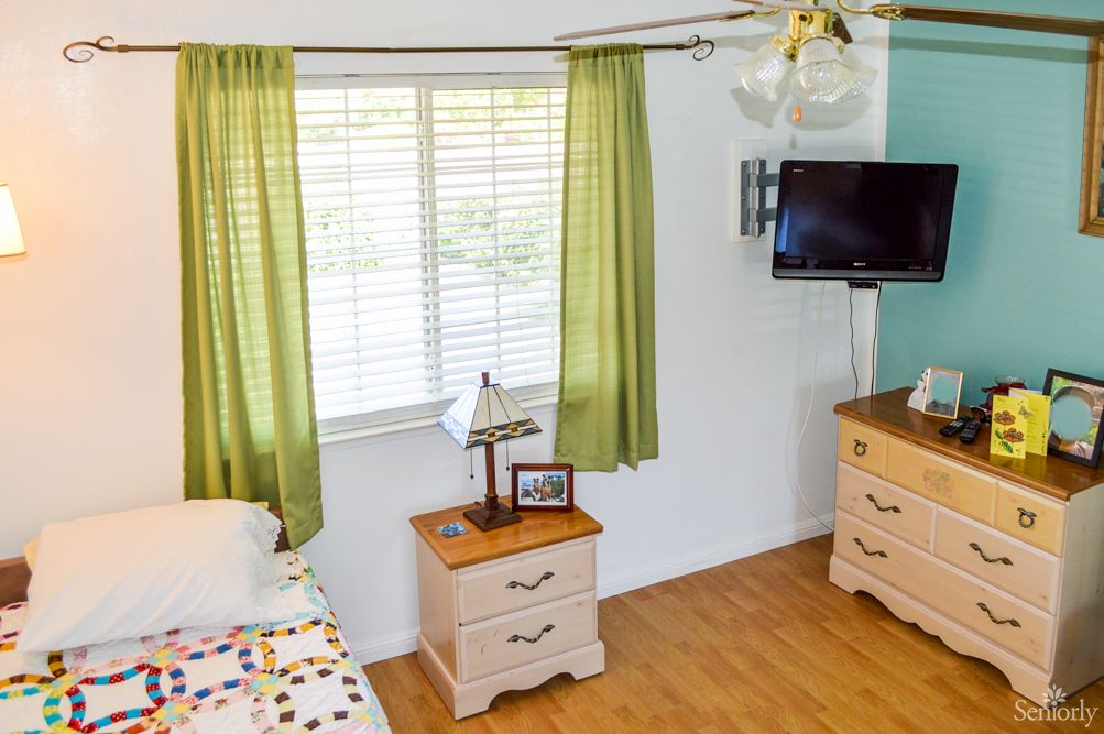 A cozy bedroom with green curtains, a nightstand with a lamp, and a wall-mounted television.
