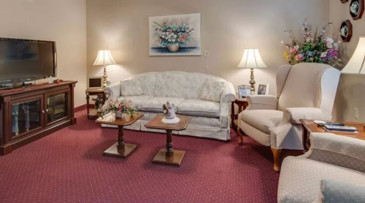 A cozy living room featuring a floral-patterned sofa, an armchair, two wooden coffee tables, and table lamps on either side. The room has a red carpet and a framed artwork on the wall.