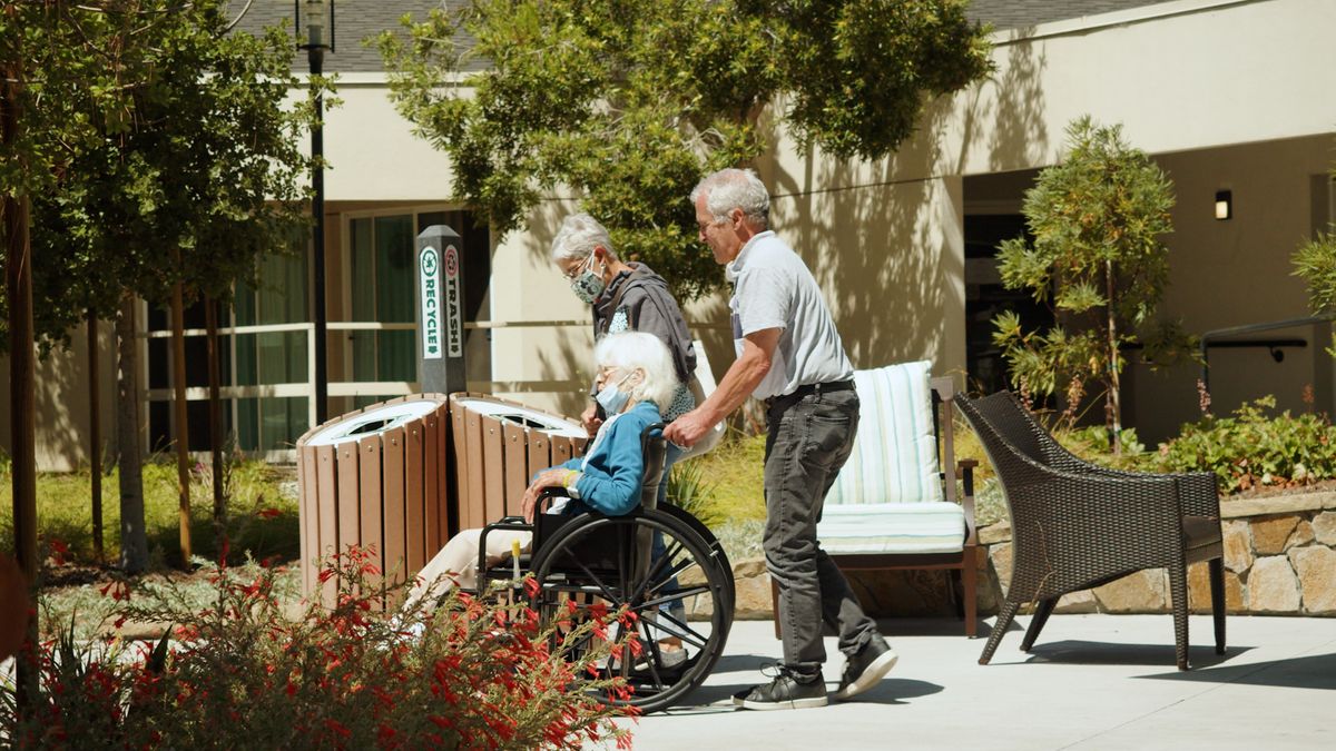 An elderly person being assisted in a wheelchair outdoors by caregivers.