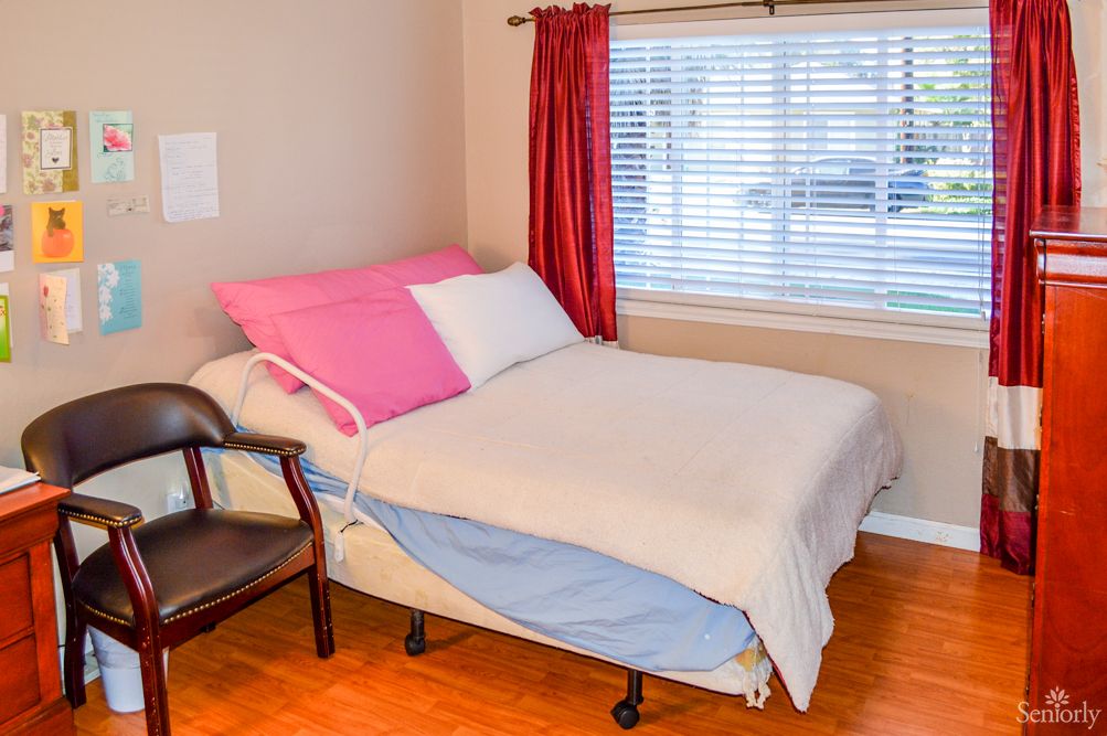 A cozy bedroom with a neatly made bed, featuring pink and white pillows, and a chair by the window with red curtains.