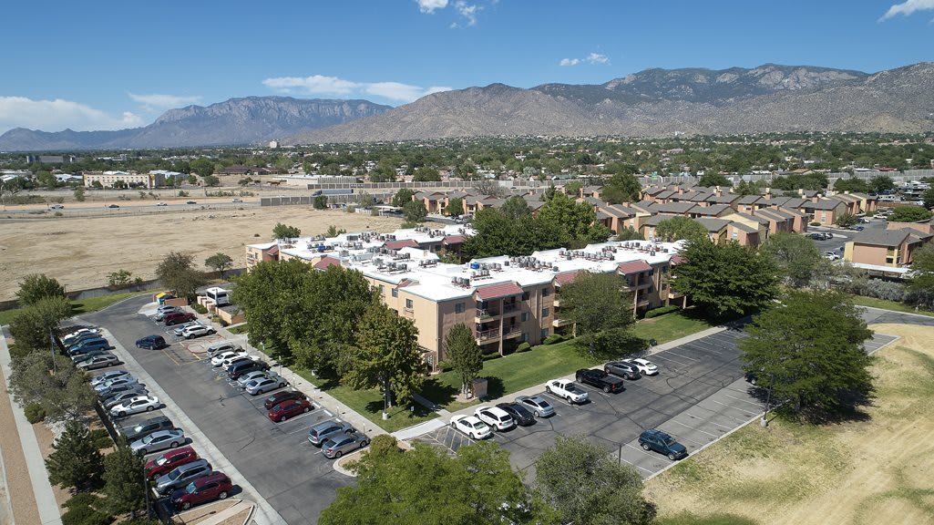 Aerial view of an apartment complex with a parking lot, surrounded by trees and mountains in the background.
