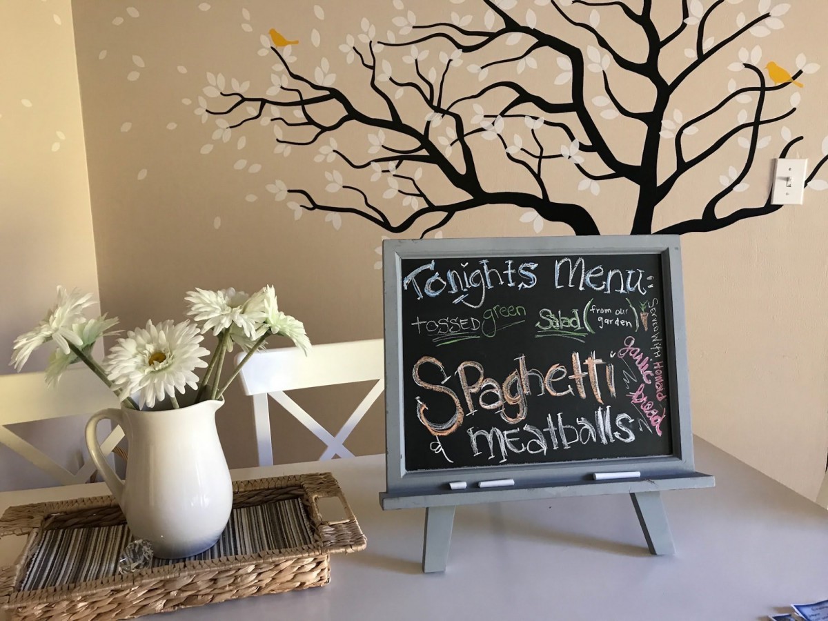 A dining room setting with a chalkboard menu displaying 'Spaghetti & Meatballs' alongside a vase of flowers and a tree mural.