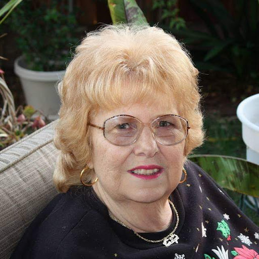 A cheerful woman sitting outdoors, wearing glasses and a festive top.