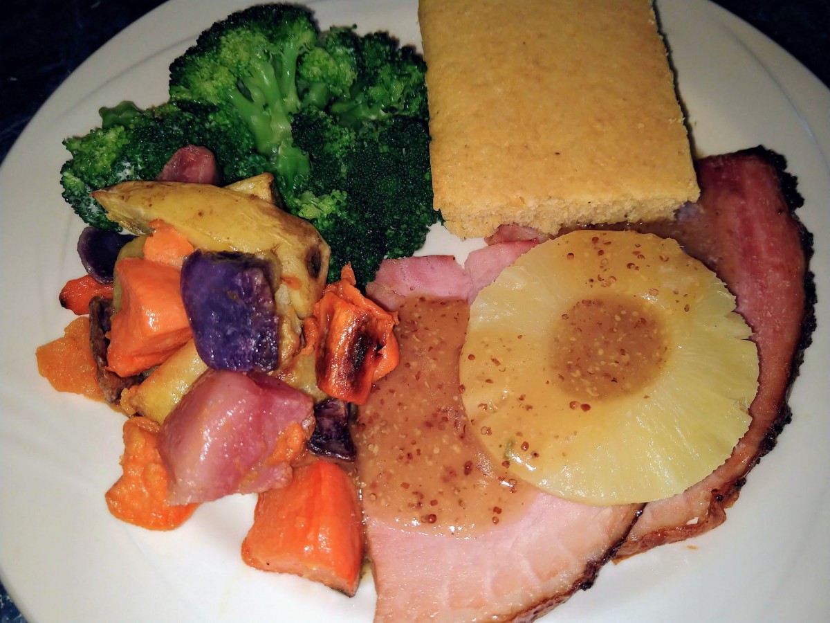 A plate featuring ham topped with pineapple, accompanied by broccoli, roasted vegetables, and a piece of cornbread.