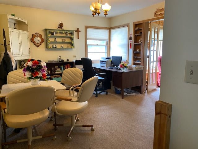 A cozy home office featuring a desk with a computer, comfortable chairs around a table, shelves with decor, and large windows providing natural light.