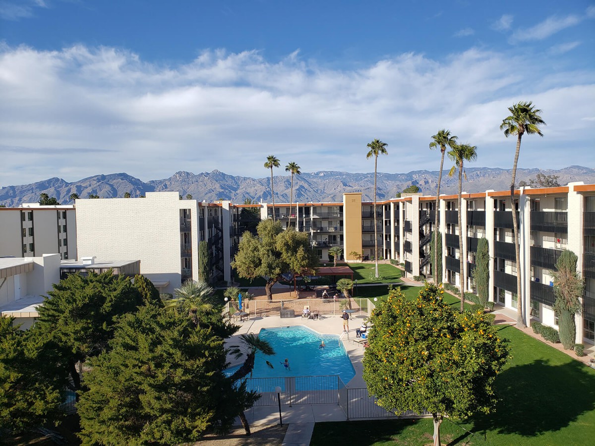 A hotel with a central swimming pool surrounded by palm trees, set against a backdrop of mountains.