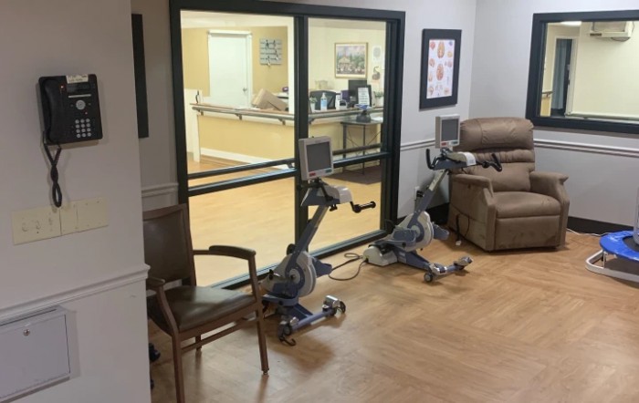 A healthcare facility room featuring two stationary bikes and chairs near a glass window.