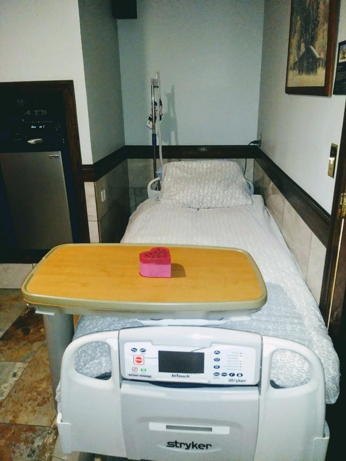A hospital bed with an IV stand in a patient room, featuring a tray table with a small pink item.