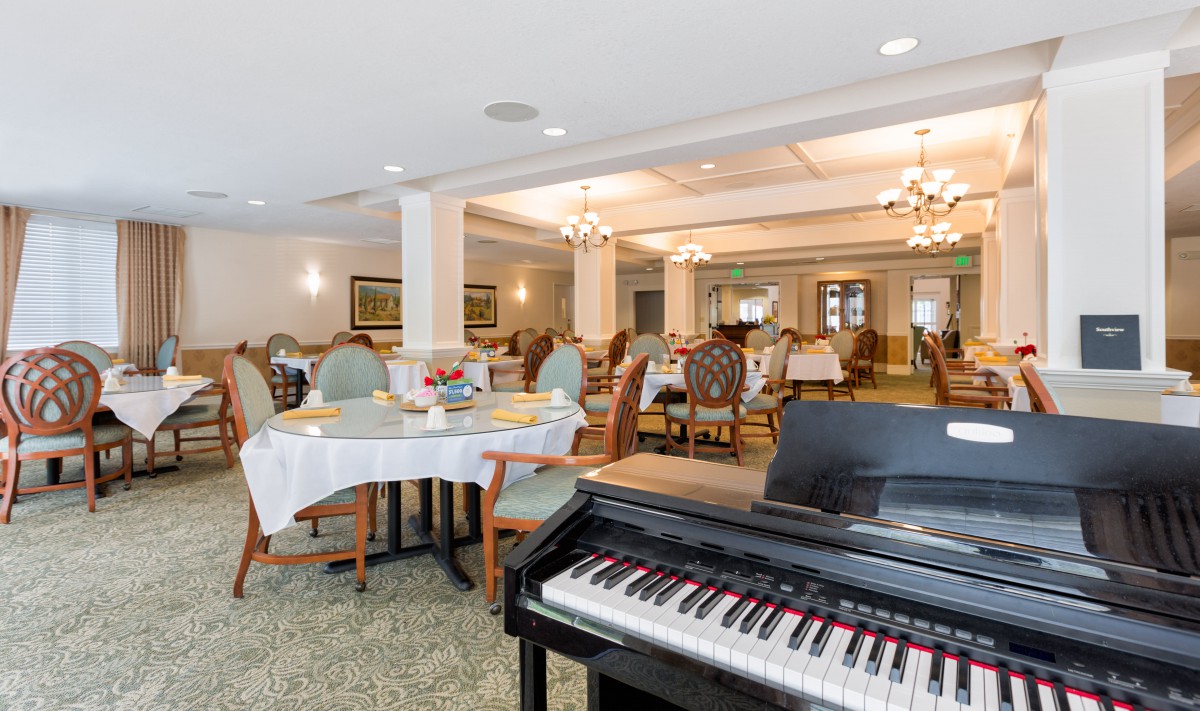 A spacious dining room with elegant decor features a piano in the foreground and several tables and chairs set for dining.