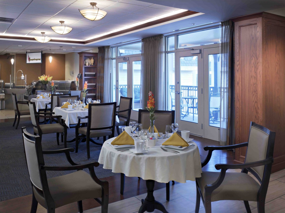 An elegant dining room with well-set tables and chairs, featuring large windows for natural lighting.