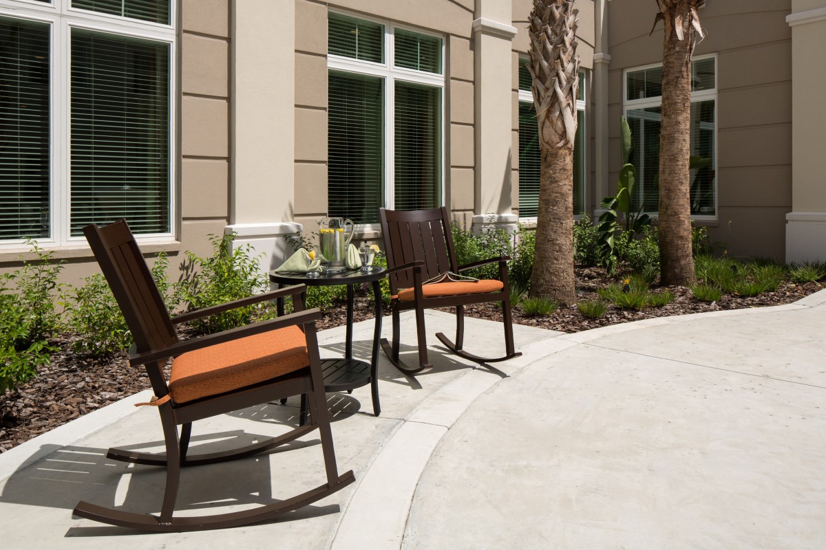 A cozy patio area with two rocking chairs and a table, surrounded by greenery and palm trees.