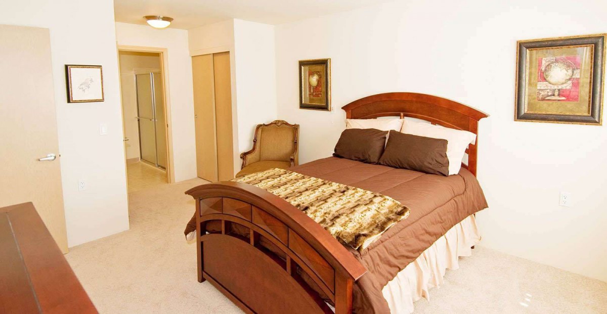 A well-lit bedroom featuring a wooden bed with brown bedding and a decorative throw, complemented by a classic chair and wall art.
