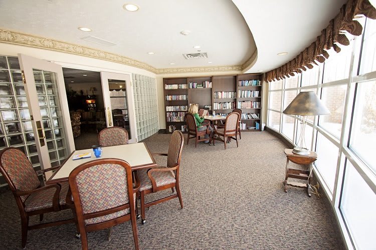 A cozy library room with bookshelves, tables, and chairs by a large window.