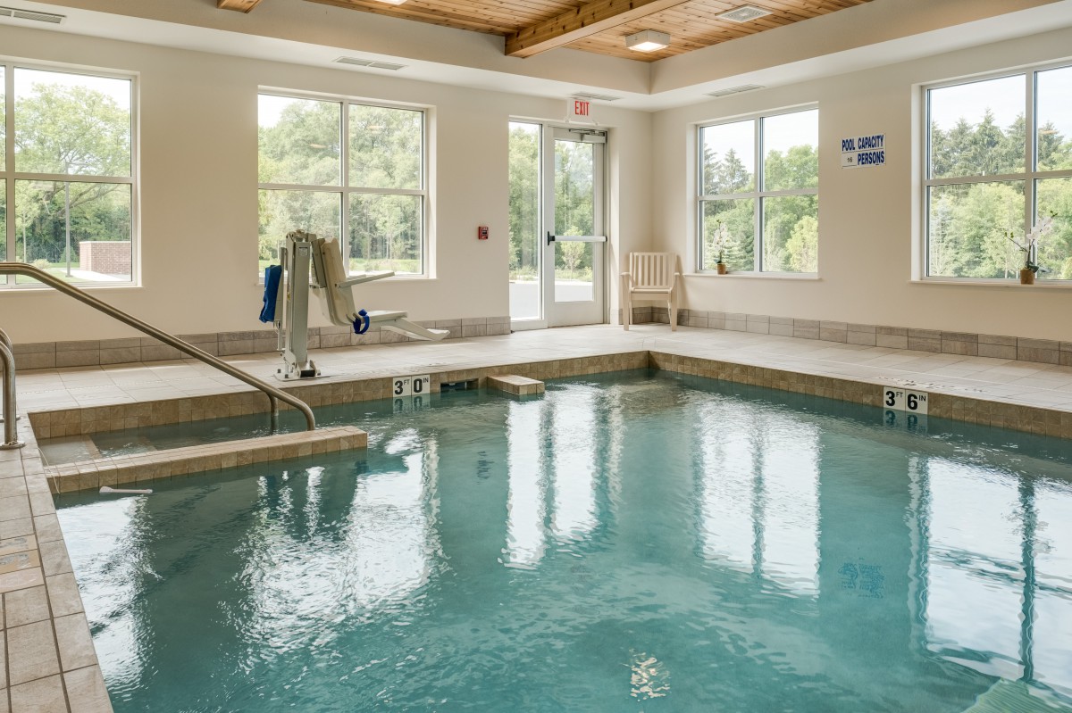 An indoor swimming pool with bright natural light streaming through the windows, featuring a pool lift for accessibility.