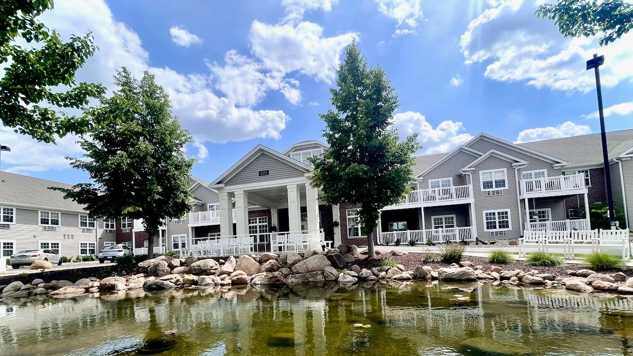 A scenic view of a large building with white columns, surrounded by trees and reflected in a tranquil pond under a partly cloudy sky.